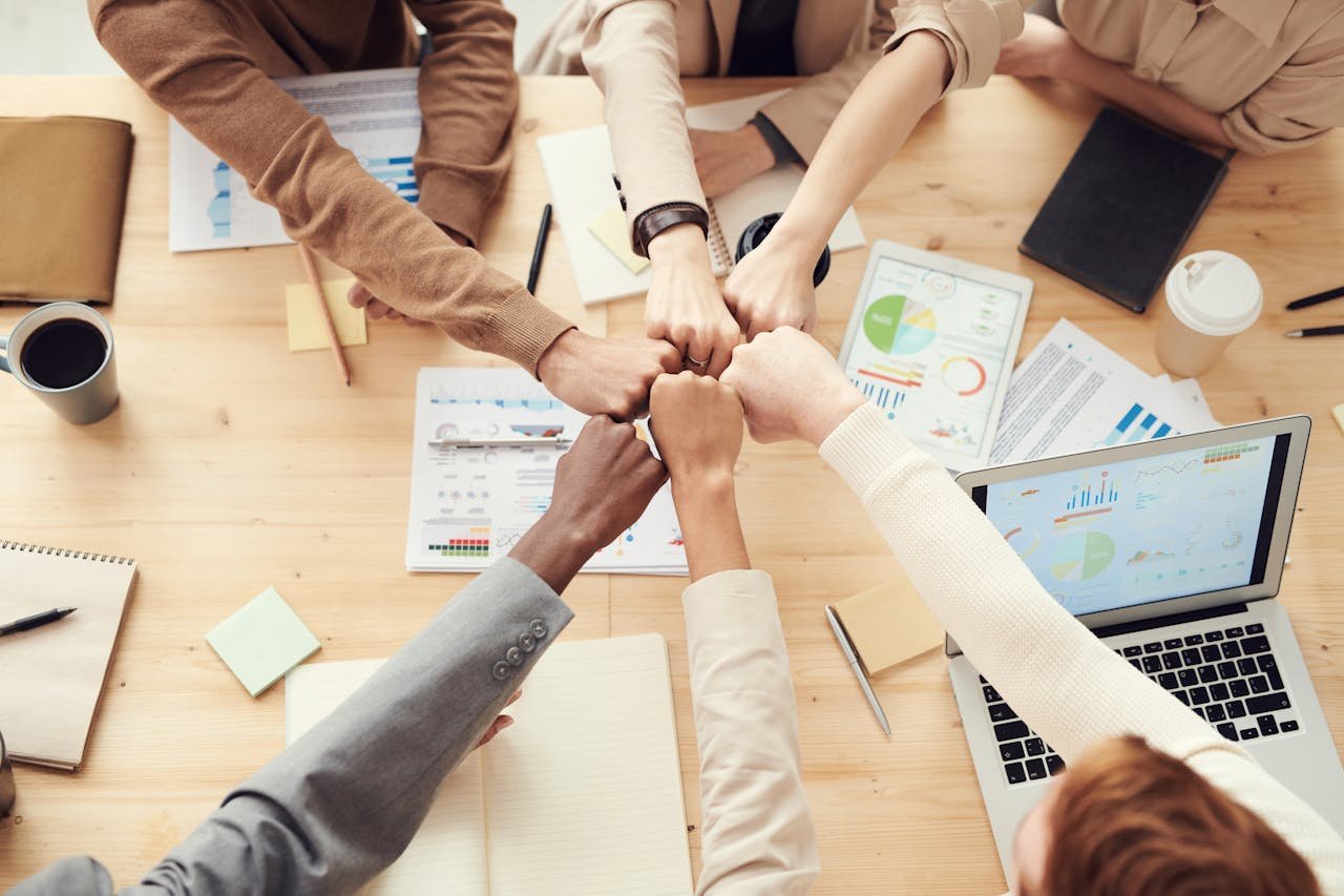 gallery-6 Top view of a diverse team fist bumping over a meeting table with paperwork and laptops, symbolizing teamwork.