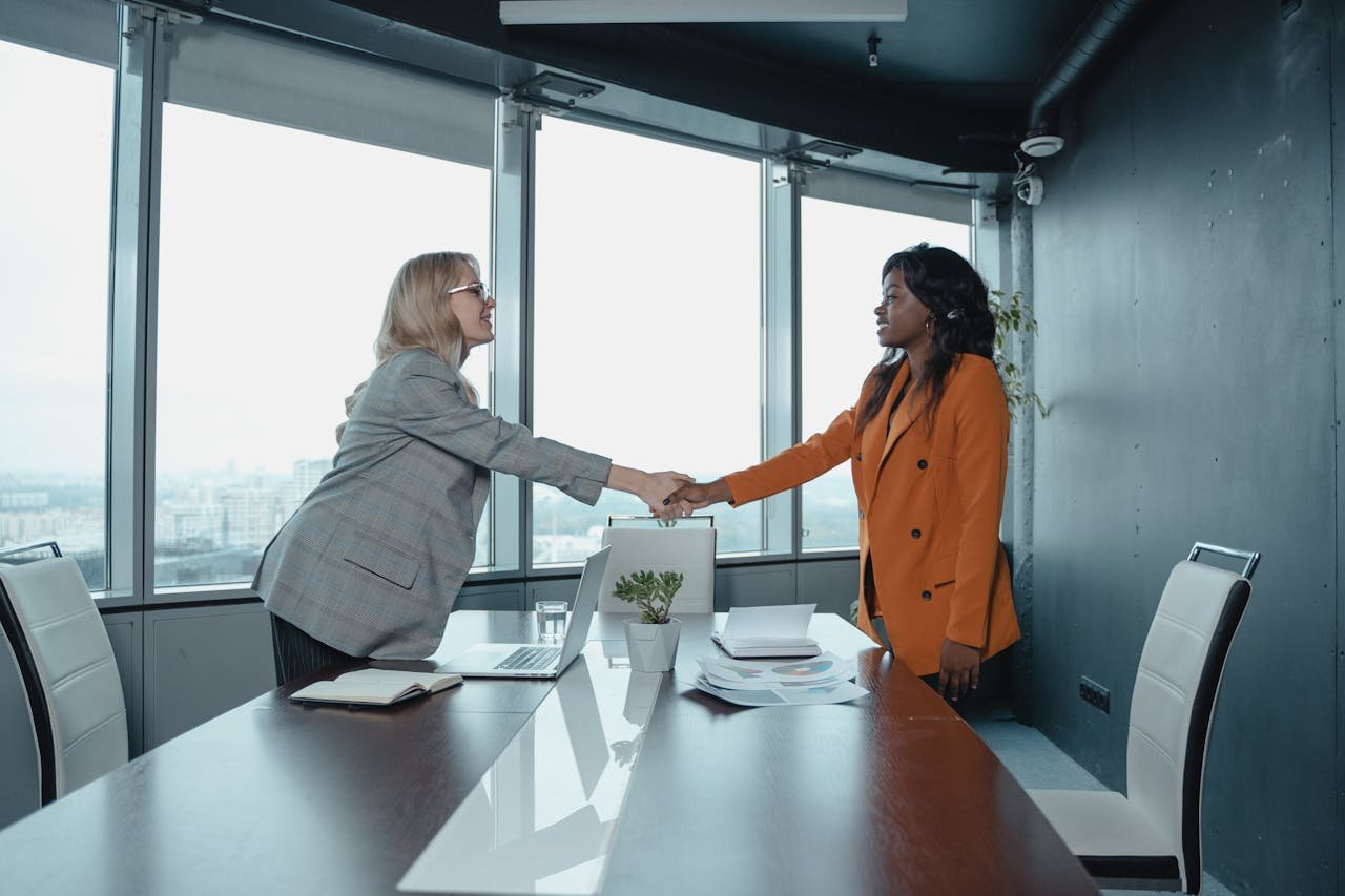 services-04 Two businesswomen shaking hands in a modern office setting, symbolizing partnership and agreement.