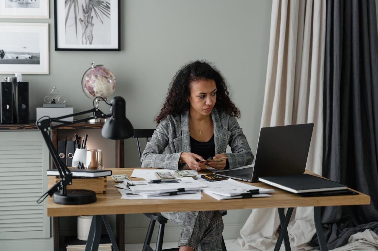 about-01 Woman in office reviewing finances with laptop, documents, and cash on table.