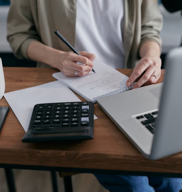 our-services-3 A woman writes financial calculations in a notebook, using a calculator and laptop at a wooden desk.